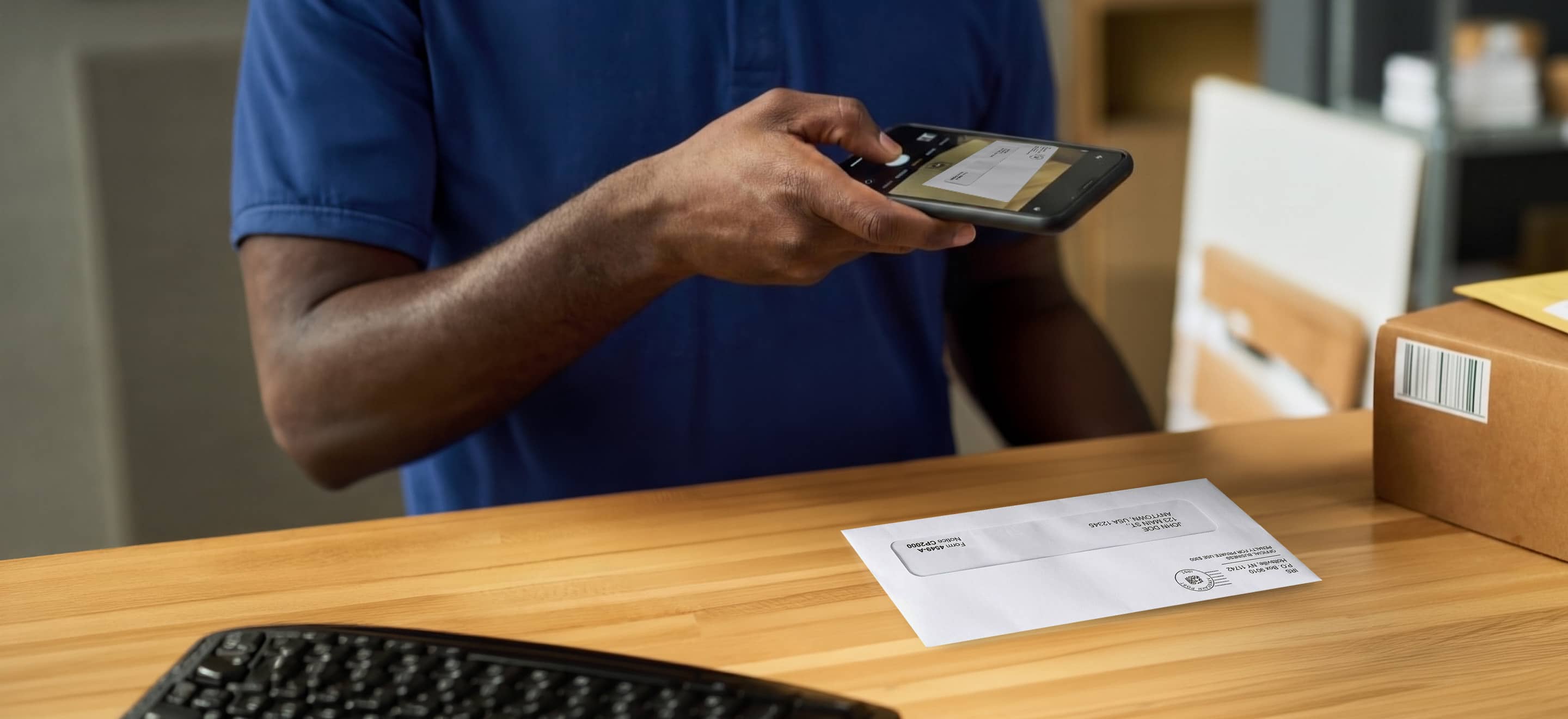 A person in a blue shirt scans a document with a smartphone on a wooden desk. Nearby are a keyboard and cardboard box. The scene is focused and professional.